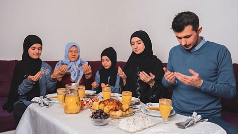 A Muslim famiily of 3 generations sit around a breakfast table, hands raised in prayer