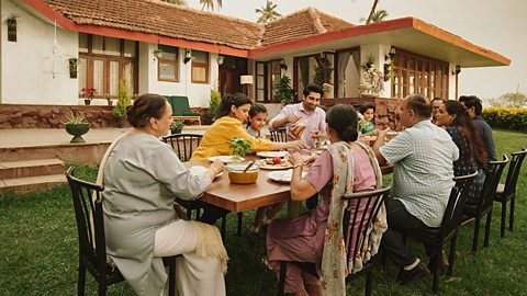 A group of people eating together at a long table on a lawn outside of a house. The people are dressed in tradition Hindu clothing and include a range of ages from Children to grandparents - A large family eating together.