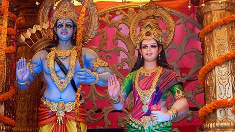 Two large statues, one a man in blue with a gold head dress and necklace, he carries a bow and arrow. The other statue is a female wearing colourful robes and a gold headpiece and necklace. Both are smiling and hold one hand up