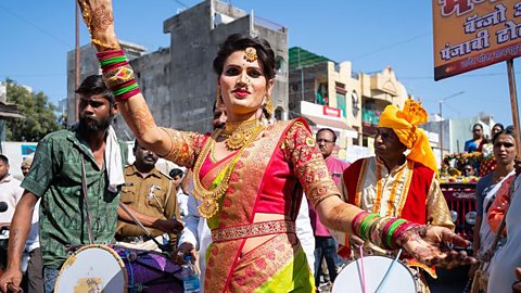 A person dressed in an elegant dress and henna on their hands dances amongst a group of men who are playing drums.