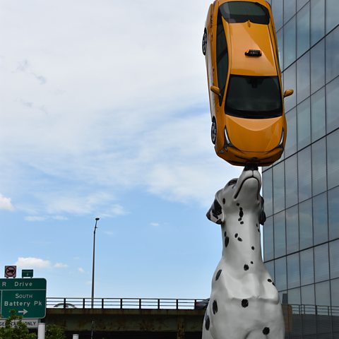 A dalmation sculpture balancing a yellow taxicab on its nose. 