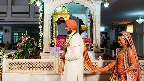 A man walks in front of a lady, the lady is holding on to the man's long decorated scarf. Both are smiling. They walk around an ornate white structure.