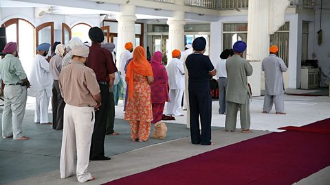 A group of male and female Sikhs stood together facing the front with their hands in prayer in a Gurdwara.