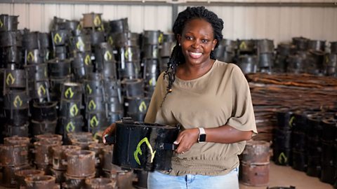 Charlot Magayi of Mukuru Clean Stoves holds one of her black stoves, with its green flame outline logo in a warehouse. There are hundreds of her stoves in the background and dozens of the old stoves her company has replaced in the foreground.