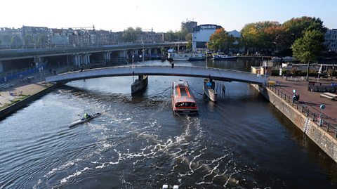 A barge crosses underneath the central section of a bridge over a canal in Amsterdam on a sunny day. There is also a single person canoe in the water. There is a series of bubbles and ripples on the surface of the water, behind the barge and in front of the canoe, that has been produced by The Great Bubble Barrier.