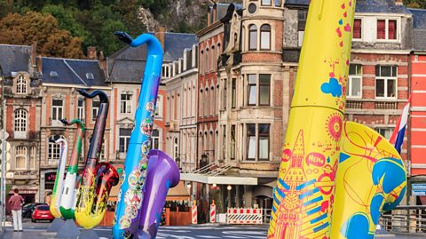 Saxophone sculptures lining the Charles de Gaulle bridge. Each of the sculptures is brightly-painted, one yellow, one blue, one black with flames, one green and one white with yellow and green stripes. Behind are some red and orange bricked buildings.