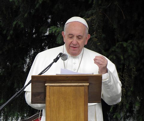 A man dressed in a white robe with a white skull cap on stands in front of a lectern with a microphone and gives a speach