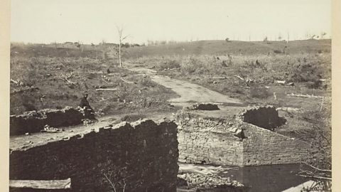 A sepia tone photo of a man sat on the edge of a ruined stone bridge, the surrounding landscape flattened and full of fallen trees