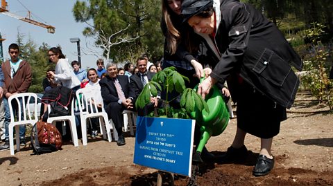 An elderly woman and her granddaughter use a watering can to water a tree sapling. A blue sign identifies its link to Anne Frank. An audience watches in white plastic chairs.