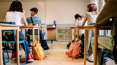 Students sat at wooden desks, looking towards the front of a classroom.