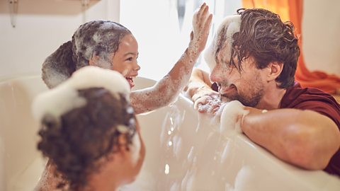 Two pre-schoolers in the bath cover their dad's face in foam