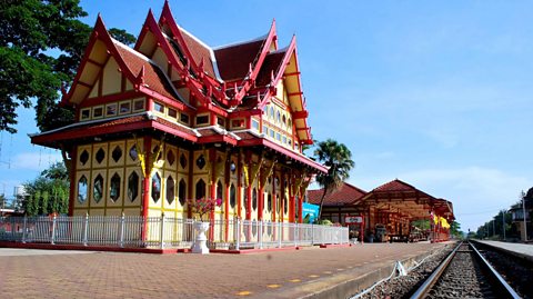 The royal pavilion of the Hua Hin Railway Station is in the foreground on the left side with the main station building in the background. The train tracks are to the right. The pavilion is red and yellow in colour and surrounded by a small metal fence.