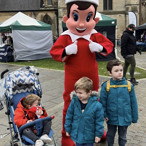 Three children standing in front of a mascot elf at a Winter market