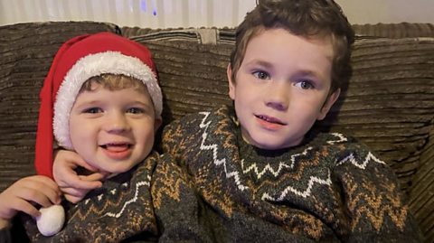 Two children in Winter jumpers. One is wearing a Santa hat.