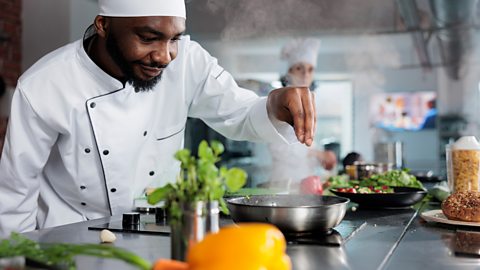 Man in chef whites seasons a pan of food on the hob, surrounded by pasta, yellow peppers and carrots.