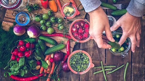 A person  putting vegetables in jars. The image is taken from the top and you can see the person's hands as well as carrots, cucumbers, radishes, peppers and onions.