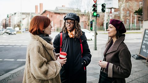 Three woman stand on a path talking to each other, one holding a coffee