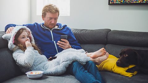 A man looks at his phone with a concerned expression, while his daughter eats snacks and watches television