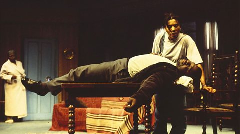 A black man lies on a table. A young black woman looks over him, whilst a middle-aged black woman stands in the background.