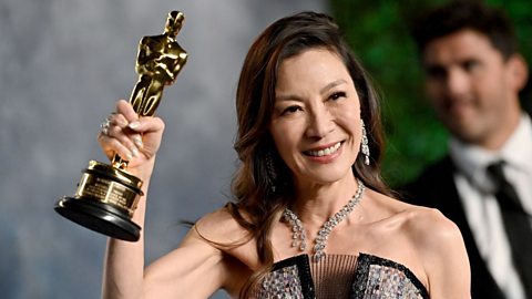 Michelle Yeoh smiles whilst holding a golden Oscar statue aloft. She wears a statement diamond necklace and earrings and a silver strapless dress.