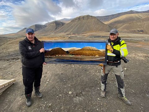 A photo of two dressed in outdoor work wear hold up a photo of a mine in front of an empty, rocky, mountainous landscape