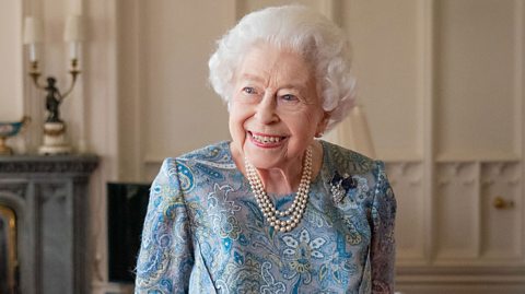 Queen Elizabeth II smiles to her right in a room in Windsor Castle. She is wearing a turquoise patterned dress and a pearl necklace.