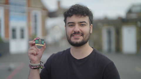 Yussef Rafik is smiling directly at the camera and holding up a micro:bit in his left hand. He is wearing a black T-shirt and the blurred background shows school buildings. 