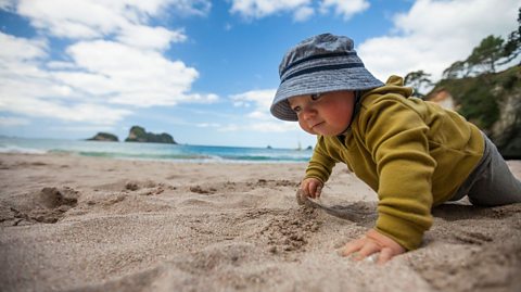 A baby crawls on the beach in Hahei, New Zealand on a bright and sunny day, with a few clouds in the sky. The baby is wearing a blue bucket hat, a yellow-green long sleeved top and grey trousers.