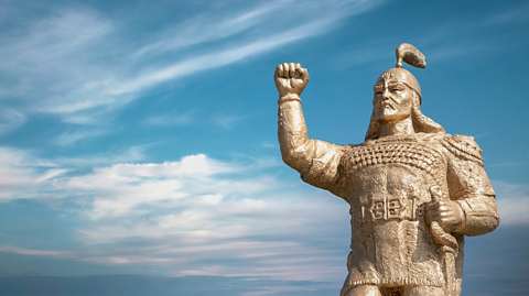 A close up image of a Manas statue against the skyline. Manas wears armour, his hand is raised. 