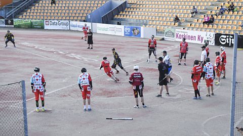 Several players for the pesäpallo team of Joensuu, wearing predominantly red kits, watch as their batter strikes the ball and attempts a run. Defending Kouvola players in black and yellow kits prepare to field the ball.