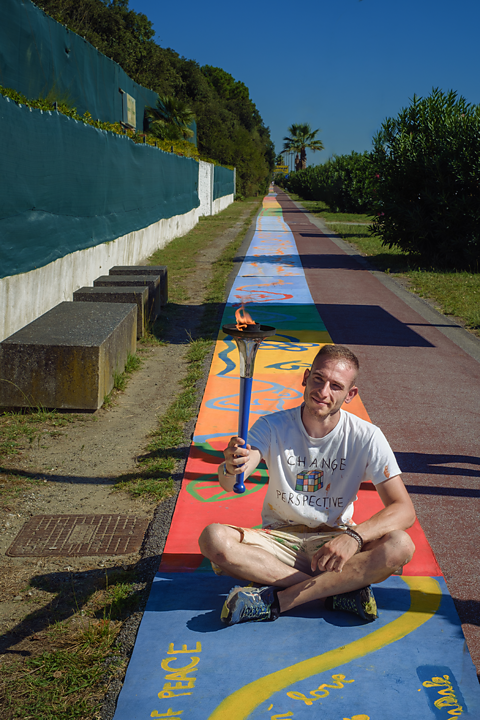 A man sits crossed legged outside on the floor. He is holding a lit torch. The floor is covered by a long, colourful artwork. 