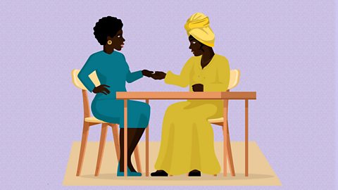 Two black women sit behind a table. One is wearing a blue dress and golden earrings. The other is wearing a long, yellow dress and head dress. They are holding hands.