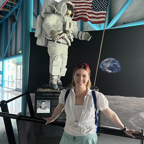 Lowena Hull is leaning against a railing in front of a space exhibit. She is smiling at the camera.