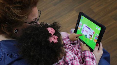 A young child sits on an adult's lap using the CBeebies Little Learners app on a tablet.