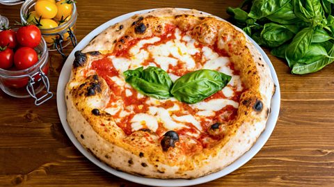 A pizza is on a plate on a wooden table. There are red and yellow tomatoes in jars to the left of the pizza and loose basil to the right. The pizza is cooked in a Neapolitan style, with a crispy, slightly charred crust and is topped with tomato sauce, cheese and two basil leafs.