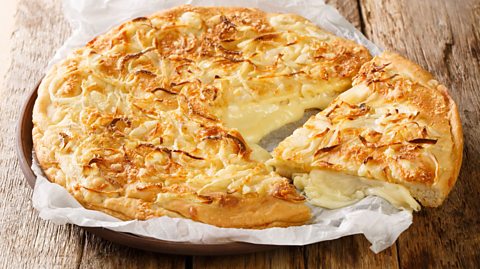 An Argentinean fugazetta pizza is served on top of greaseproof paper on top of a plate, on a wooden table. It is a pizza with two layers of dough, with a thick cheesy layer in between. Browned onions are on top of the top layer. A slice has been pulled to reveal the oozy cheese centre.