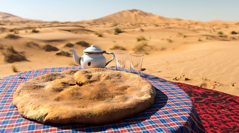 A madfouna is served on a gingham placemat on a table. A teapot and two glass cups are behind it. In the distance, the sands of the Saharan desert can be seen.