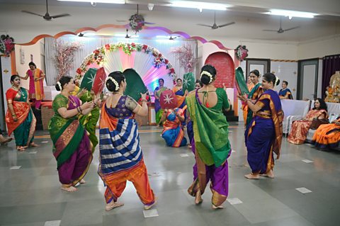 A group of women dressed in colourful silk robes dance and clap in a hall.