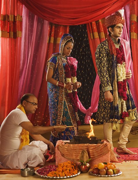 A Hindu bride and groom dressed in patterned silks with gold detailing stand next to a small sacred fire with their scarves tied together. A priest in plain pale clothes sits by the fire adding offerings to it.