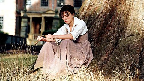 An actress playing Elizabeth Bennet. She is wearing a white shirt and pale pink dress, and is sat on the grass beneath a tree.