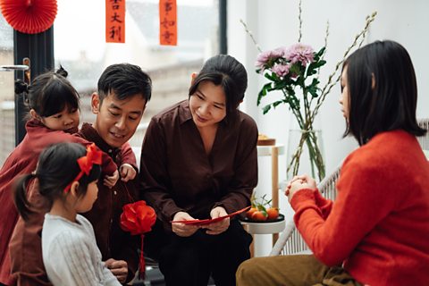 A young Asian family are gathered together and smiling. The mother passes a red envelope to a small girl.