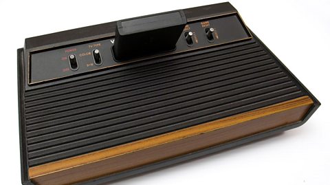 A shot of the Atari Video Computer System, later renamed to the Atari 2600. It is a black rectangular console, with four levers that control, from left to right, the power, whether it's a colour or black and white television, game selection and resetting the game. In between levers two and three is a black game cartridge. The front wall of the console has a wood veneer panel.