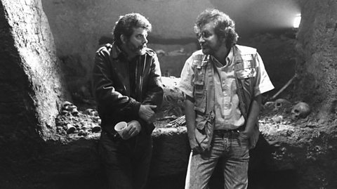 George Lucas and Steven Spielberg are sat together in a cave-like structure, used on the set of Indiana Jones and the Last Crusade. Lucas is on the left, holding his arm and a coffee cup while Spielberg has one hand in his pocket.