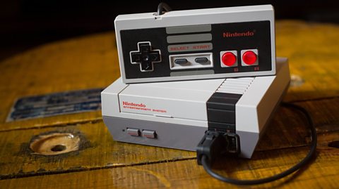 A mini Nintendo Entertainment System is sat on a wooden table. One controller is plugged in and sat atop of the console, with the buttons facing the camera.