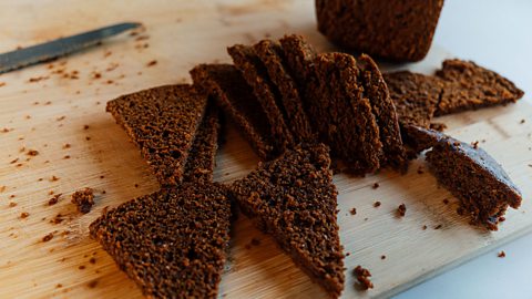 A dark brown bread sliced into triangles on a wooden chopping board with crumbs and a breadknife in the background.