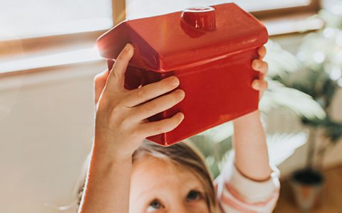 A young girl holds out a red model of a house, looking upwards at it.