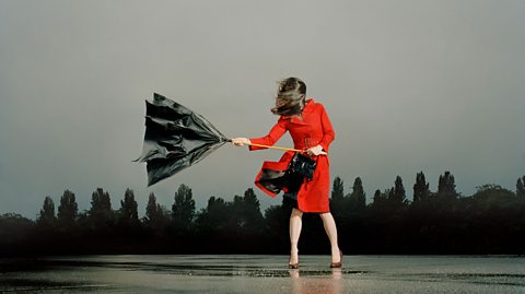 A woman wearing a red dress, holding an umbrella which is getting blown inside out by the wind.