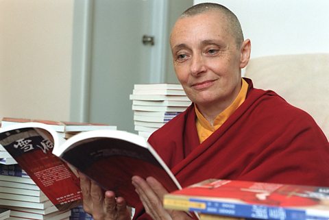 Jetsunma Tenzin Palmo sat in a room, reading a book, surrounded by multiple stacks of books.