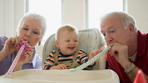 Two grandparents blow party horns for their baby grandchild, who sits in a high chair.