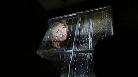 Silhouettes of people in the dark who are watching the last episode of Friends. On screen is a blond woman looking out the window as it rains.